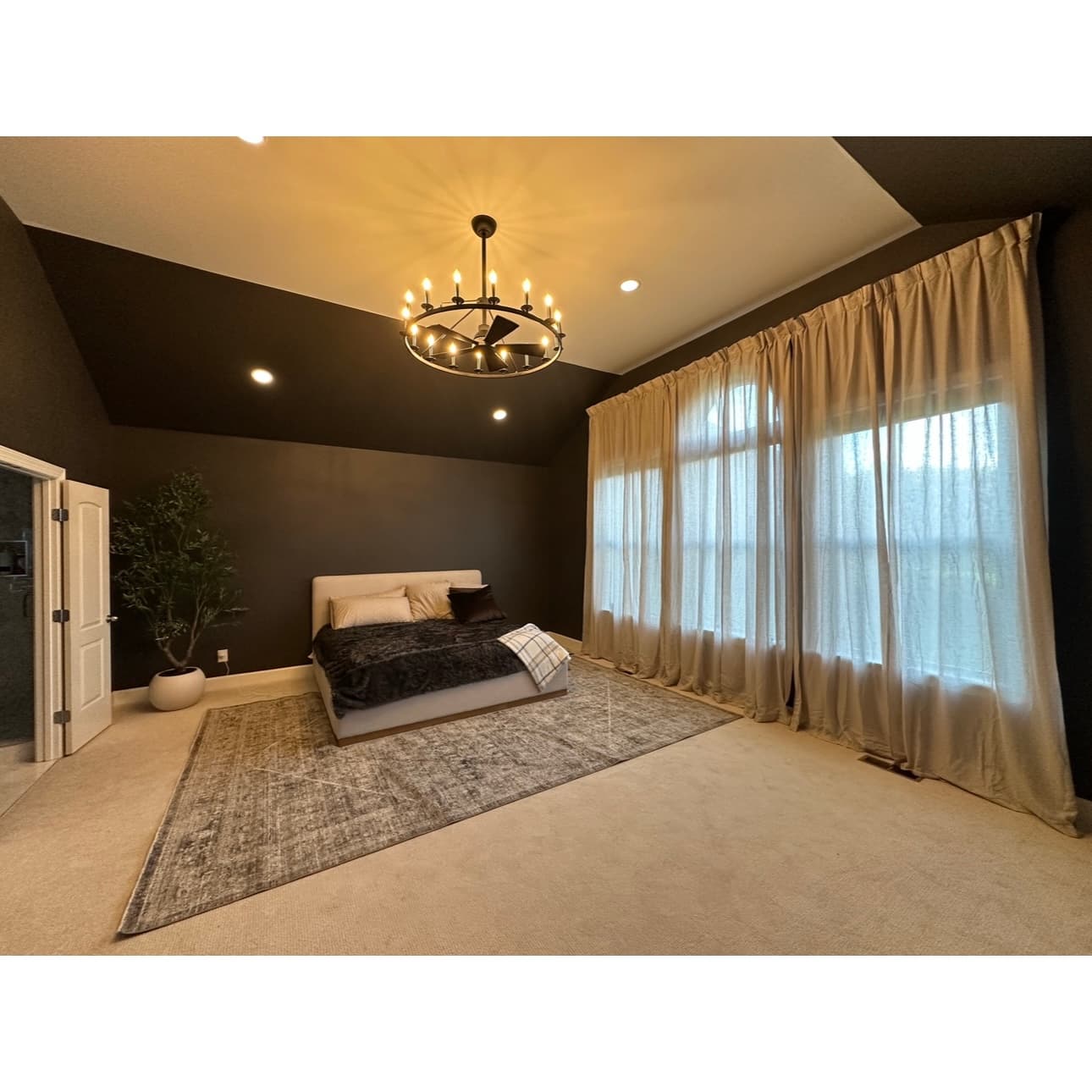 Black ring chandelier installed on a vaulted ceiling in a primary bedroom, with full-height curtains hung across the window wall