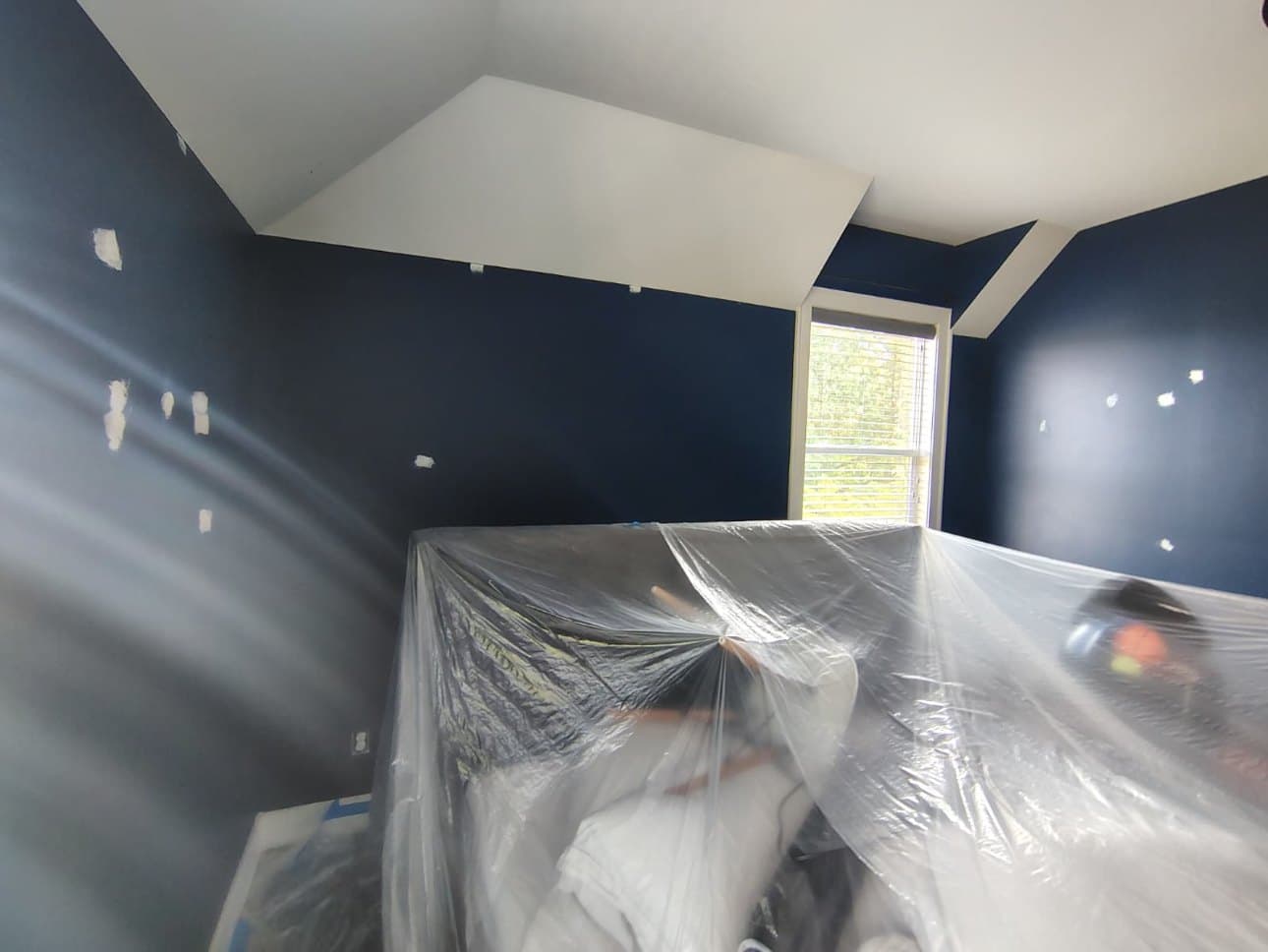 Bedroom mid-job: walls in deep navy with pale filler patches still showing, furniture sheeted in plastic